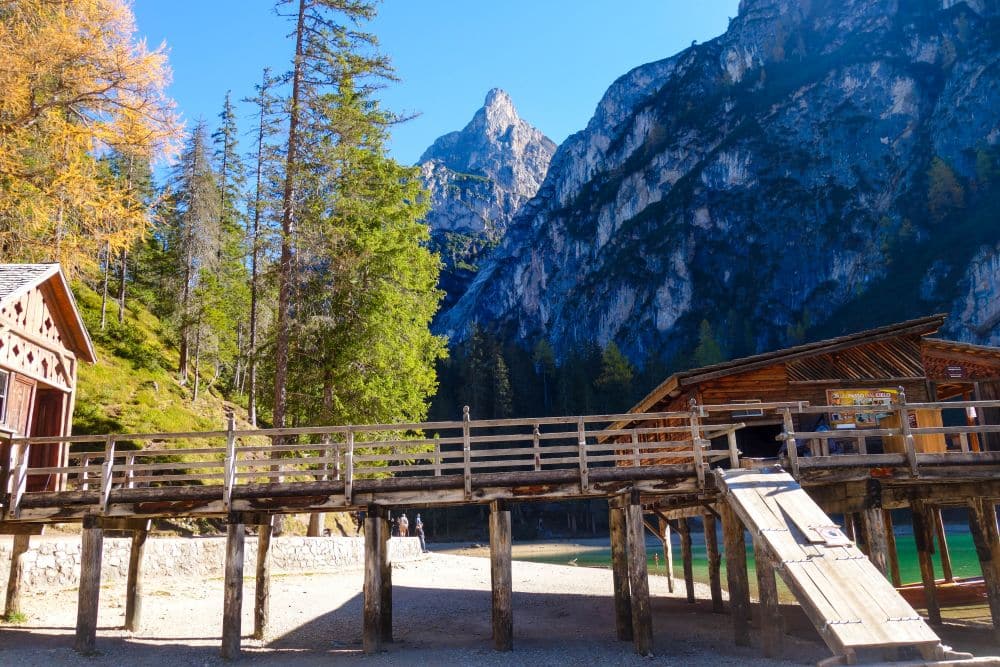 Holzhütten und ein erhöhter Steg am klaren Pragser Wildsee, umgeben von hohen Tannen und felsigen Bergen unter einem strahlend blauen Himmel.