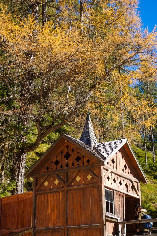 Ein kleiner, hölzerner, kapellenartiger Bau mit spitzem Dach steht im Sonnenlicht unter hohen gelben Herbstbäumen am Pragser Wildsee, vor einem klaren blauen Himmel. Zwei Personen sitzen am Rande des Gebäudes.