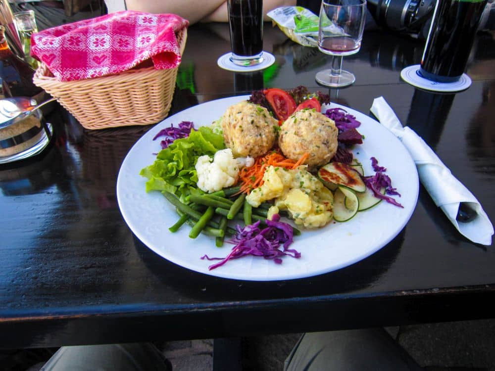 Ein Teller mit zwei Semmelknödeln, verschiedenen Gemüsesorten, Kartoffelsalat und lila Kraut steht auf einem dunklen Tisch am Brandiswaalweg, daneben ein Brotkorb, Getränke, ein Weinglas und ein in eine Serviette eingewickeltes Besteck.