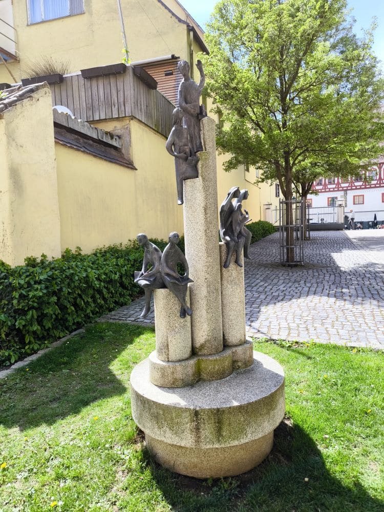 Eine Skulptur in Nördlingen zeigt vier menschliche Figuren, die auf hohen, zylindrischen Steinsäulen klettern und sitzen. Sie stehen auf einem runden Sockel auf einer Wiese in der Nähe gelber Gebäude und eines Baumes.