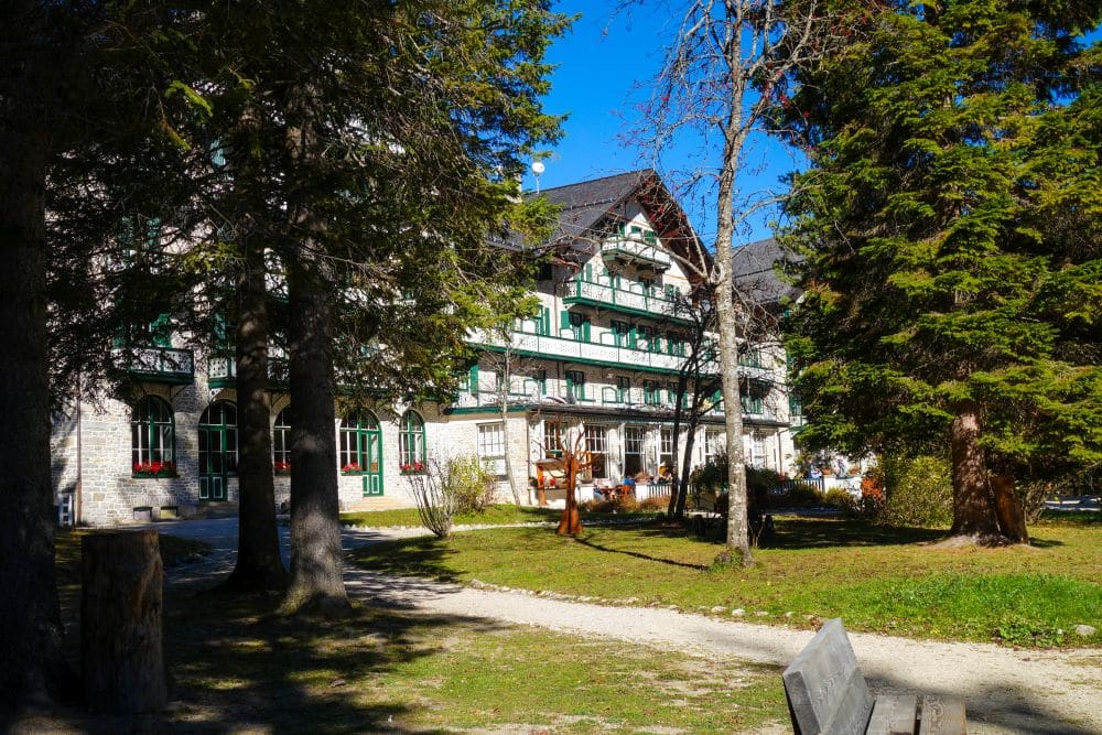 Großes Gebäude im alpenländischen Stil mit grünen Verkleidungen und Balkonen, in der Nähe des Pragser Wildsees, umgeben von hohen Immergrünen und einem sonnenbeschienenen Garten, mit einer Holzbank und unbefestigten Wegen im Vordergrund unter einem klaren blauen Himmel.