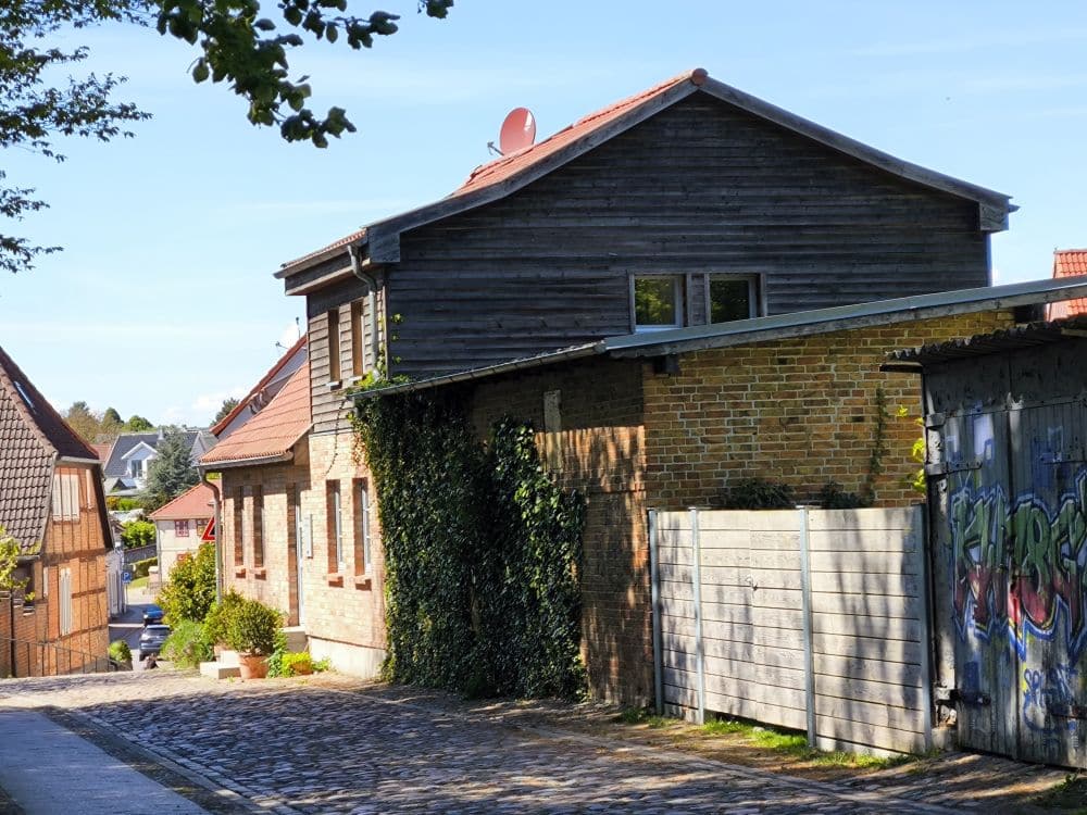 Eine ruhige Kopfsteinpflasterstraße in Klütz, in der Nähe der Ostseeküste, gesäumt von Backsteinhäusern - eines mit Holzverkleidung und einer Ranke an der Wand -, einem Holzzaun und einem mit Graffiti-Kunstwerken verzierten Garagentor unter einem klaren blauen Himmel.