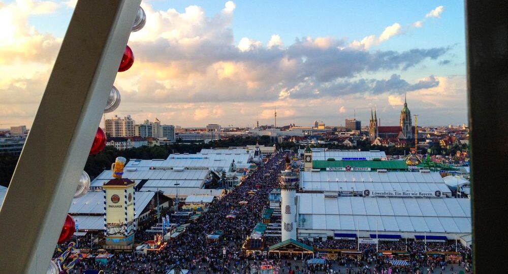 Luftaufnahme eines überfüllten Oktoberfestes mit großen weißen Zelten, bunten Ständen und städtischen Gebäuden im Hintergrund unter einem teilweise bewölkten Himmel, gesehen durch die Struktur eines Riesenrads.