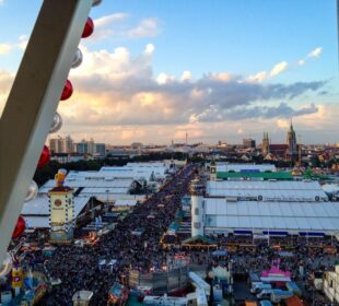 Luftaufnahme eines überfüllten Oktoberfestes mit großen weißen Zelten, bunten Ständen und städtischen Gebäuden im Hintergrund unter einem teilweise bewölkten Himmel, gesehen durch die Struktur eines Riesenrads.