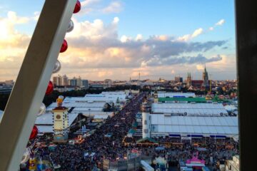 Luftaufnahme eines überfüllten Oktoberfestes mit großen weißen Zelten, bunten Ständen und städtischen Gebäuden im Hintergrund unter einem teilweise bewölkten Himmel, gesehen durch die Struktur eines Riesenrads.