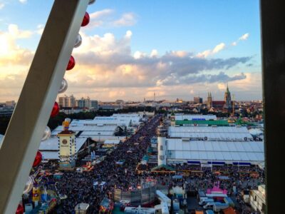 Luftaufnahme eines überfüllten Oktoberfestes mit großen weißen Zelten, bunten Ständen und städtischen Gebäuden im Hintergrund unter einem teilweise bewölkten Himmel, gesehen durch die Struktur eines Riesenrads.