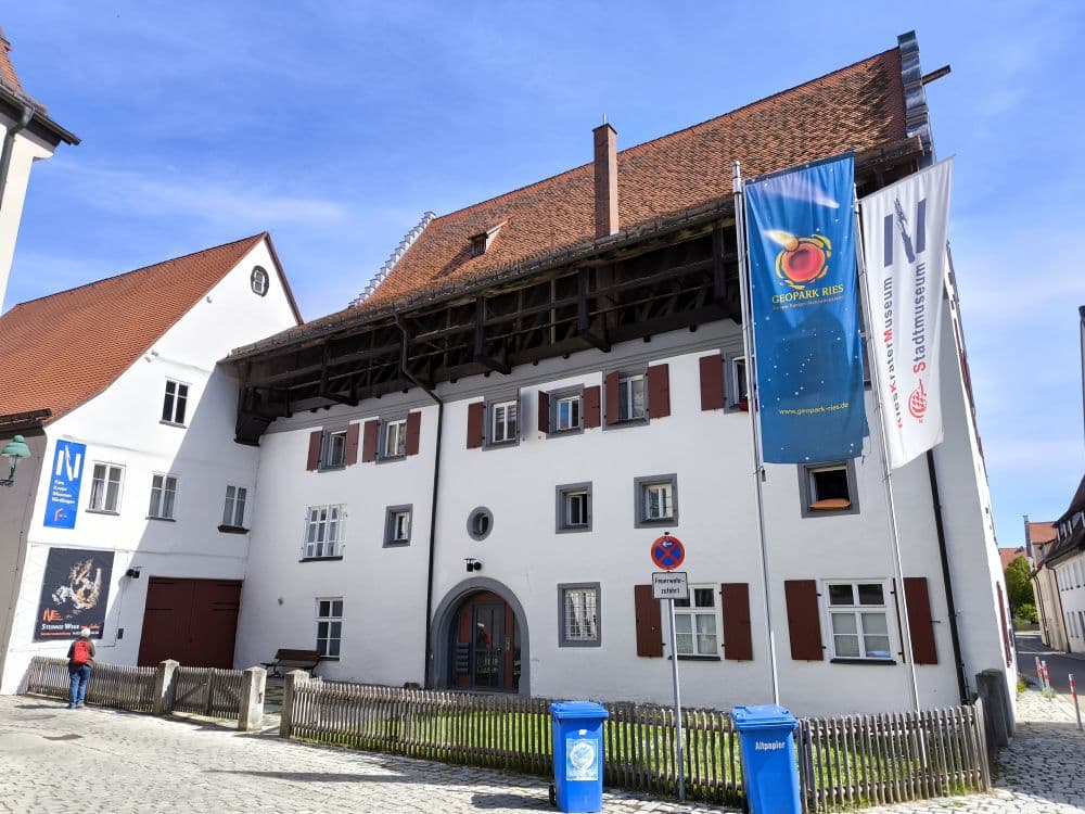 Ein historisches weißes Gebäude mit roten Fensterläden und einem steilen Ziegeldach steht in einer Kopfsteinpflasterstraße in Nördlingen. Vor dem Haus wehen blau-weiße Fahnen, und an einem Holzzaun in der charmanten Stadt auf der Schwäbischen Alb stehen Mülltonnen.