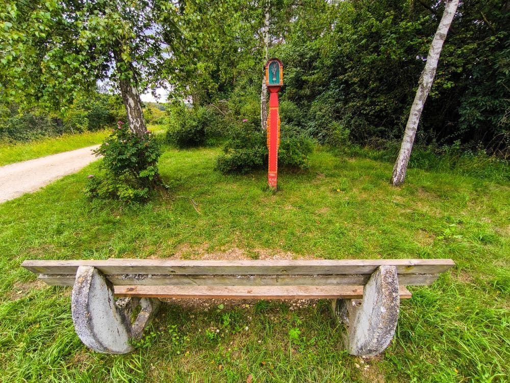 Eine Holzbank steht im Gras vor einem kleinen roten Schrein, umgeben von Grün und Bäumen im Hintergrund in der Nähe von Hayingen; ein Feldweg, Teil der Hochgehschätzt Wanderung Glastal, windet sich nach links.
