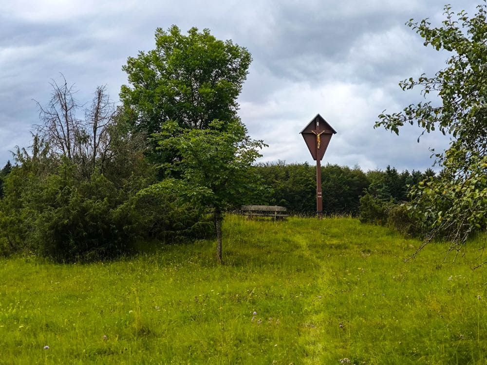 Ein Holzkreuz auf einem hohen Pfosten steht auf einer Wiese bei Hayingen, umgeben von grünen Bäumen und Sträuchern unter einem wolkenverhangenen Himmel - eine einladende Kulisse für eine Wanderung durch das malerische Glastal. Hinter den Bäumen ist teilweise eine Holzbank zu erkennen.