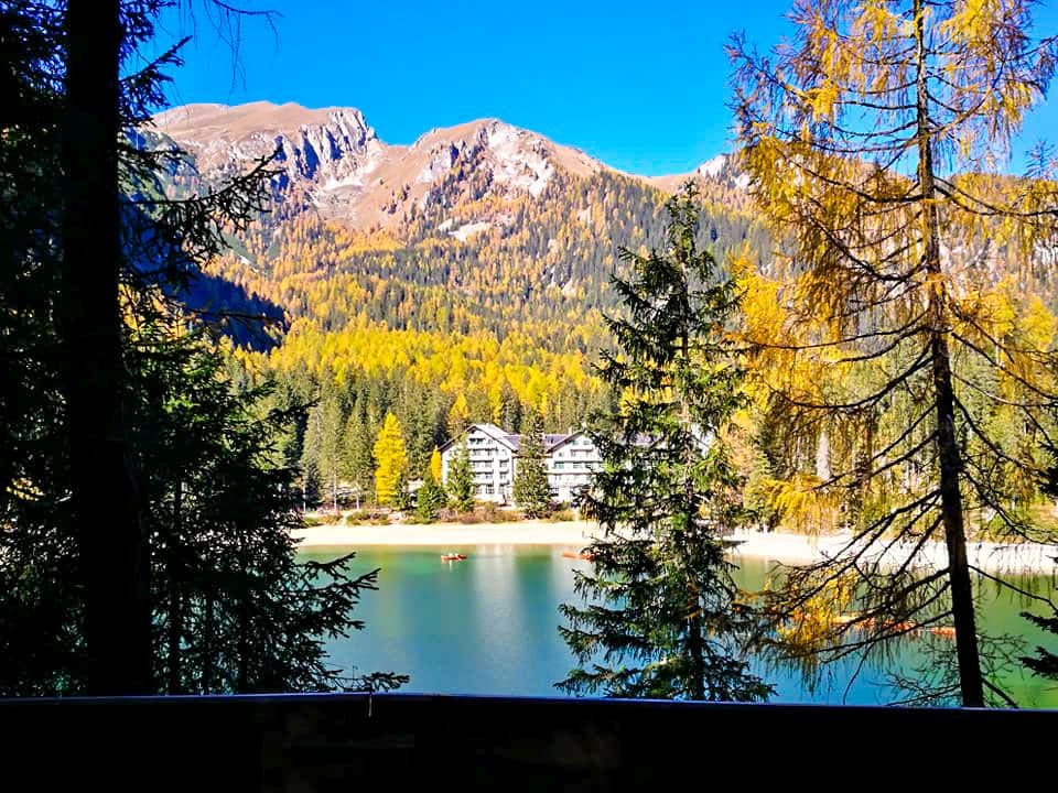 Ein malerischer Blick auf den Pragser Wildsee, einen von immergrünen und herbstlichen Bäumen umgebenen See, mit Bergen im Hintergrund und einigen Gebäuden in der Nähe des Seeufers unter strahlend blauem Himmel.