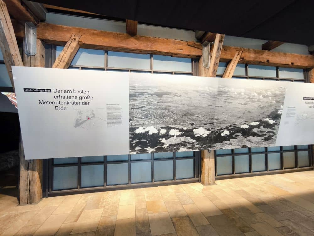 Eine große Ausstellungstafel im Rieskratermuseum zeigt eine Luftaufnahme und einen deutschen Text über das Nördlinger Ries, einen der besterhaltenen Meteoritenkrater der Erde, der an einer Wand mit Holzbalken und Fenstern angebracht ist.