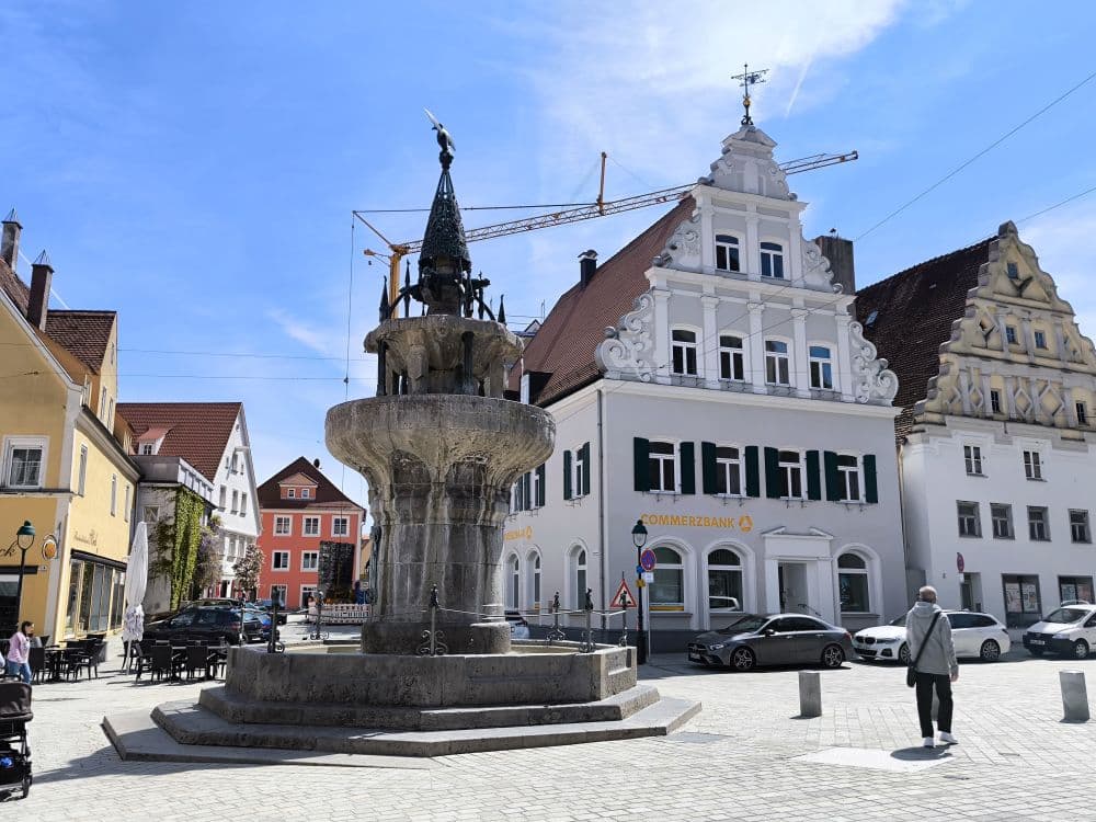 Ein Steinbrunnen steht auf einem Stadtplatz in Nördlingen, umgeben von historischen Gebäuden mit Giebeldächern. Eine Person geht in der Nähe spazieren, und im Hintergrund ist bei teilweise bewölktem blauen Himmel ein Kran zu sehen.