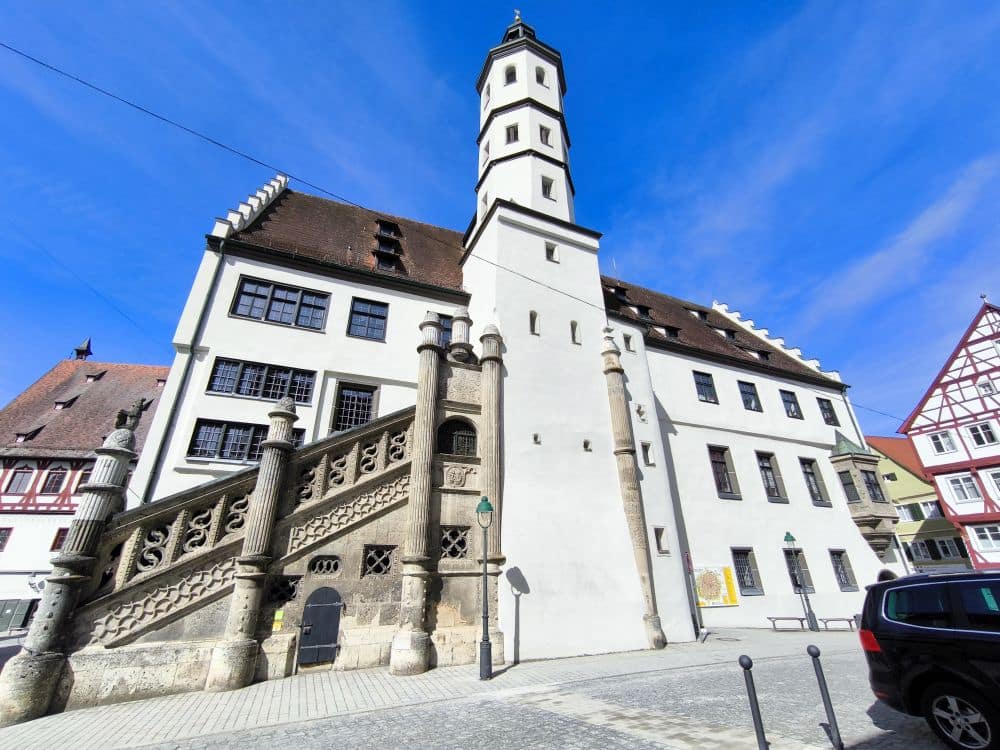 Historisches weißes Gebäude mit einem hohen zentralen Turm, einer verzierten Steintreppe und einem schrägen rotbraunen Dach unter einem strahlend blauen Himmel. In Nördlingen gelegen, säumen nahe gelegene Gebäude und parkende Autos die charmante Kopfsteinpflasterstraße.