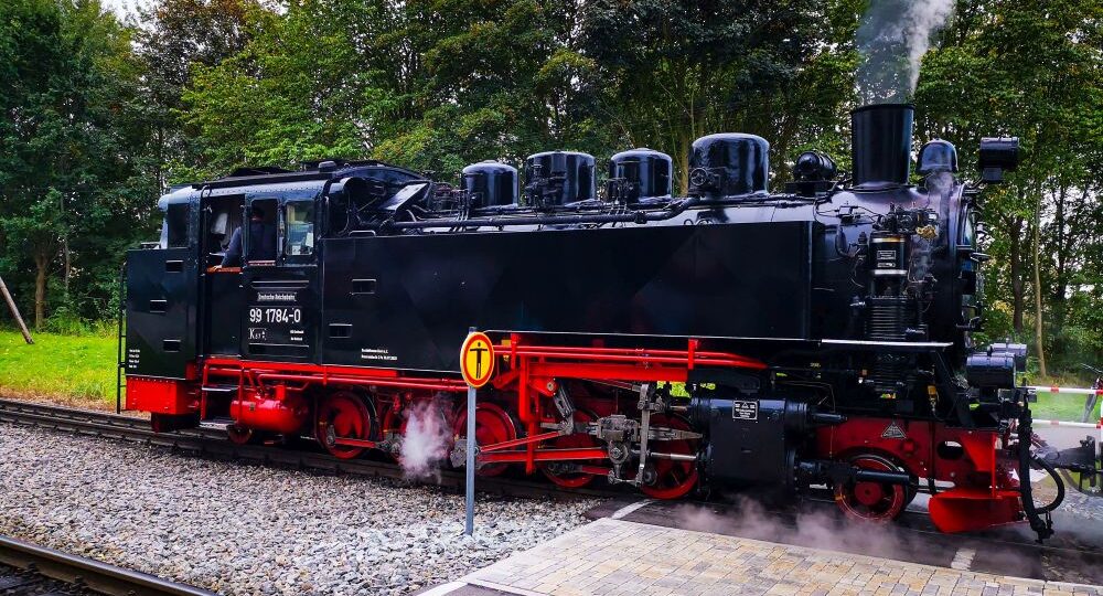 Eine alte schwarze Dampflokomotive, bekannt als Rasende Roland, mit roten Rädern und Details stößt Dampf aus, während sie auf den Bahngleisen in der Nähe einer gepflasterten Fläche, umgeben von Bäumen und Grün auf der Insel Rügen, steht.