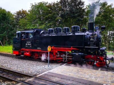Eine alte schwarze Dampflokomotive, bekannt als Rasende Roland, mit roten Rädern und Details stößt Dampf aus, während sie auf den Bahngleisen in der Nähe einer gepflasterten Fläche, umgeben von Bäumen und Grün auf der Insel Rügen, steht.