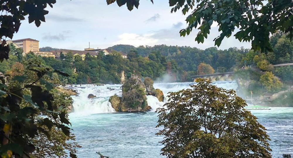 Ein breiter Wasserfall, der Rheinfall bei Schaffhausen, fließt über Felsen, umgeben von Bäumen mit Herbstlaub, mit einem Gebäude und bewaldeten Hügeln im Hintergrund unter einem bewölkten Himmel. Erleben Sie Europas größten Wasserfall in atemberaubender Kulisse.