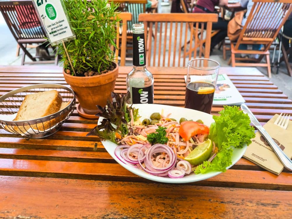 In einem Freiluftcafé in Hayingen stehen auf einem Holztisch ein frischer Salat, ein Brotkorb, ein Kräutertopf, Getränke und ein Flaschengetränk - perfekt, um nach einer Wanderung im Glastal zu entspannen. Holzstühle tragen zur gemütlichen Hochgehschätzt-Atmosphäre bei.
