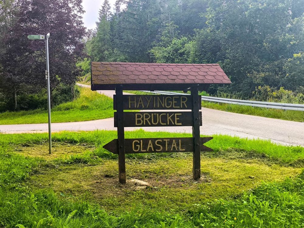 Ein hölzerner Wegweiser mit Schindeldach steht im Gras und weist auf die Hayinger Brücke und das Glastal hin - perfekt für Ihr nächstes Hochgehschätzt oder Ihre Wanderung Glastal. Im Hintergrund sind Bäume und eine Straßenkurve zu sehen.