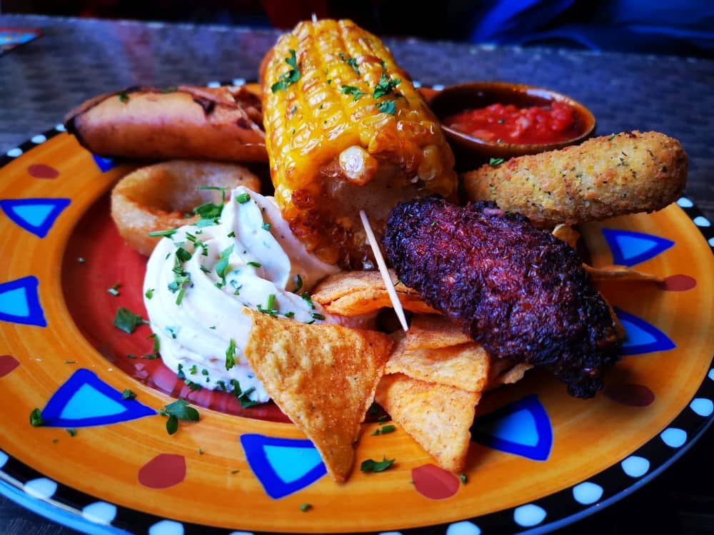 Ein bunter Teller, inspiriert von den Lieblingsrestaurants auf Rügen: gegrillte Maiskolben, Brotstäbchen, Kartoffelpüree, Nacho-Chips, Chicken Wing, Mozzarella-Stäbchen, Zwiebelring und eine rote Dip-Soße - perfekt zum Genießen auf der Insel Rügen.