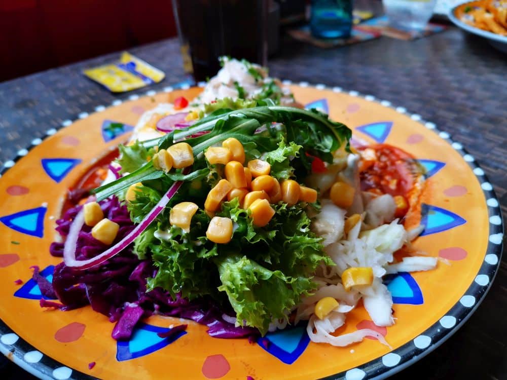 Ein farbenfroher Salat mit Kopfsalat, Mais, Kohlrabi, roten Zwiebeln und gemischtem Gemüse wird auf einem leuchtend gelben Teller mit dekorativem Rand serviert - perfekt, um ihn in einem Ihrer Lieblingsrestaurants auf der Insel Rügen zu genießen.