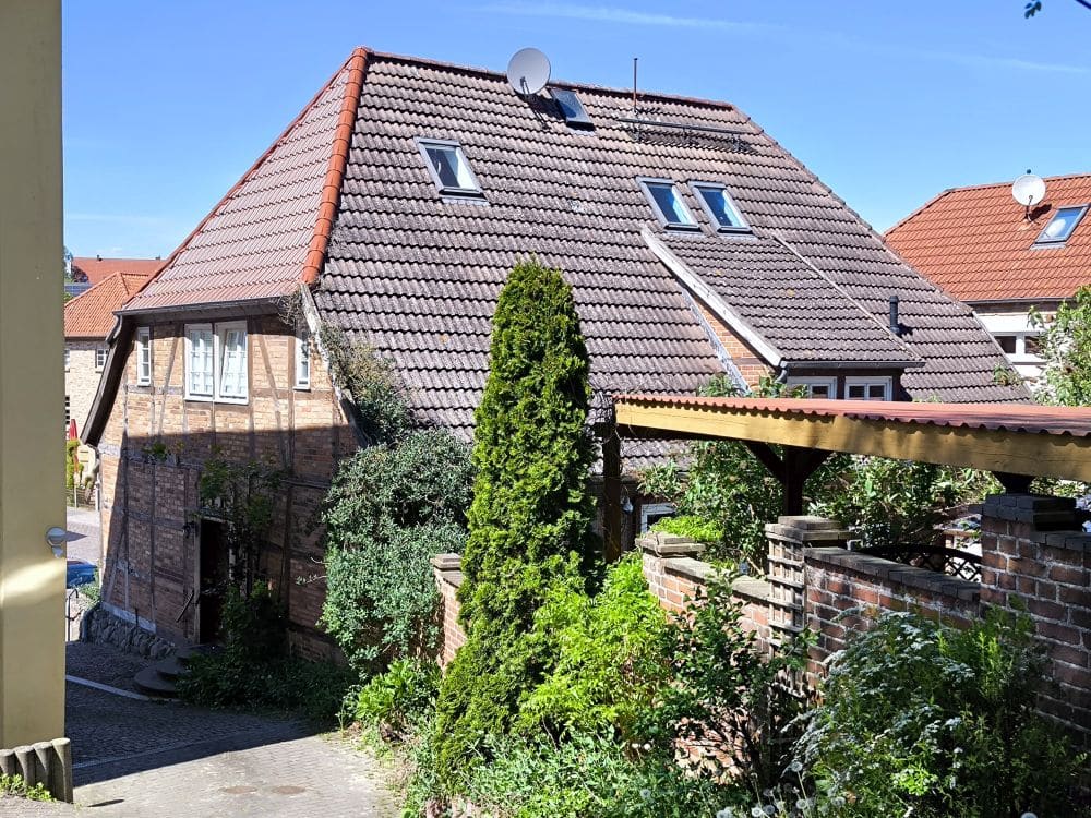 Ein zweistöckiges Haus mit rotem Ziegeldach und Dachgauben steht hinter einer Backsteinmauer, umgeben von Grün bei Klütz an der Ostseeküste. Rechts ist unter dem strahlend blauen Himmel ein Carport mit einem Metalldach zu sehen.