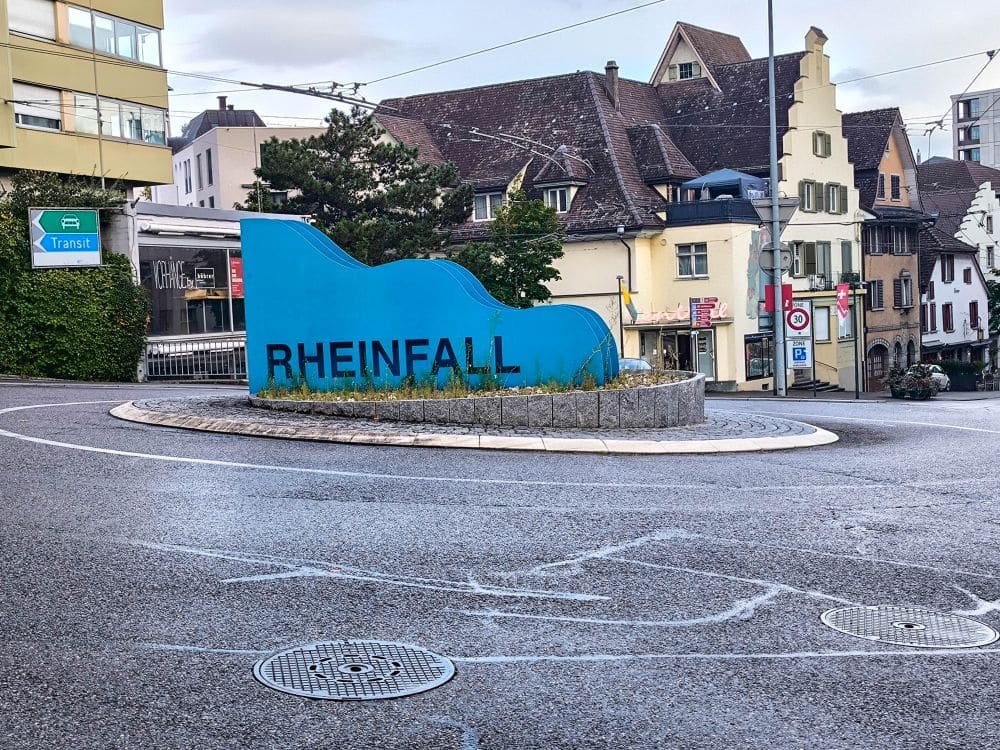 Eine blaue Skulptur in Form einer Welle mit dem Schriftzug "RHEINFALL" steht in der Mitte eines Kreisverkehrs in Schaffhausen, umgeben von Gebäuden mit traditioneller europäischer Architektur, und feiert den größten Wasserfall Europas.