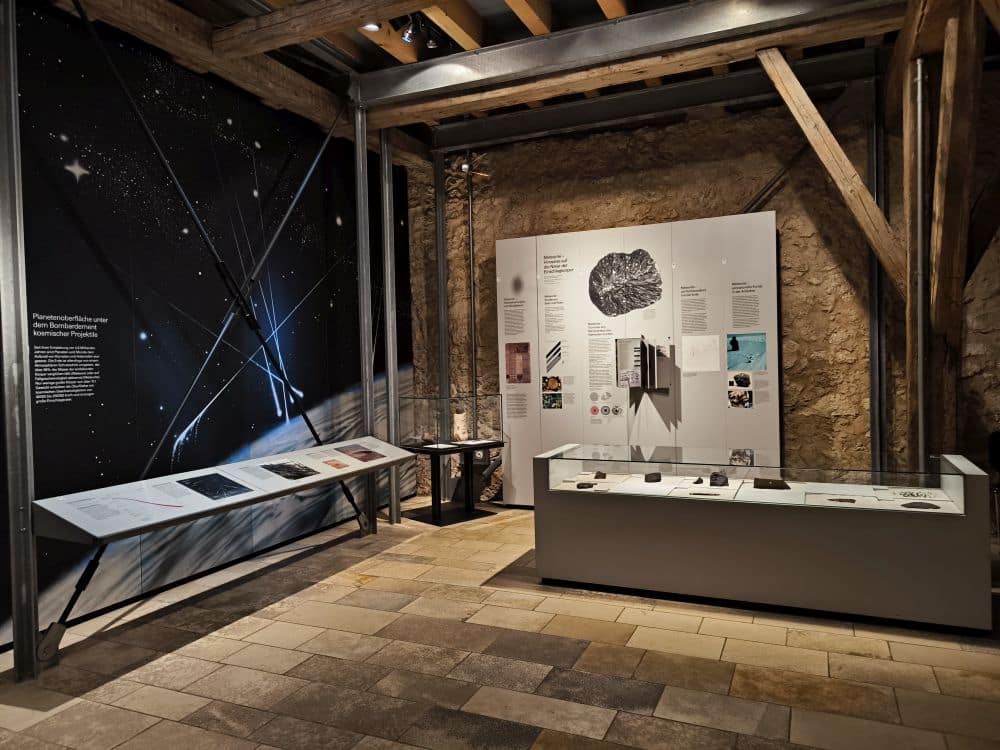 Museumsexponat im Rieskratermuseum in Nördlingen mit Steinwänden und Holzbalken, die Schautafeln zum Thema Weltraum oder Astronomie enthalten. Die Tafeln zeigen Texte, Bilder und Meteoritenartefakte. Die linke Tafel zeigt einen Sternenhimmel über der Schwäbischen Alb.