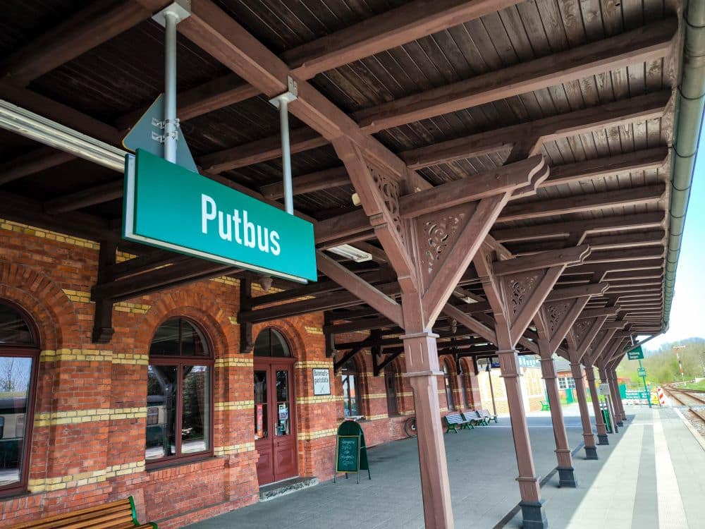 Ein überdachter Gehweg mit Holzbalken am Bahnhof Putbus auf Rügen. Der Bahnhof hat rote Backsteinwände, Rundbogenfenster, Bänke und ein grünes Schild mit Putbus in weißen Buchstaben. Hier hält der Rasende Roland in echter Volldampf-Manier.