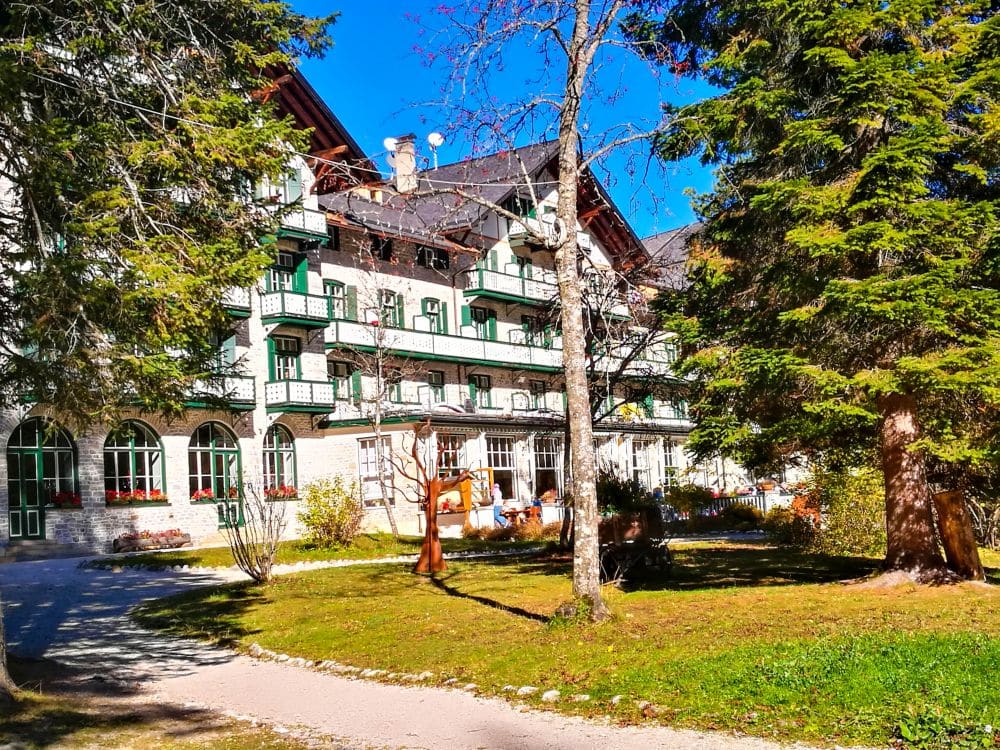 Ein großes, charmantes Gebäude in der Nähe des Pragser Wildsees mit grünen Balkonen und weißen Wänden liegt an einem sonnigen Tag hinter hohen Bäumen und einer Wiese. Vor dem Gebäude schlängelt sich ein Weg durch den Garten.