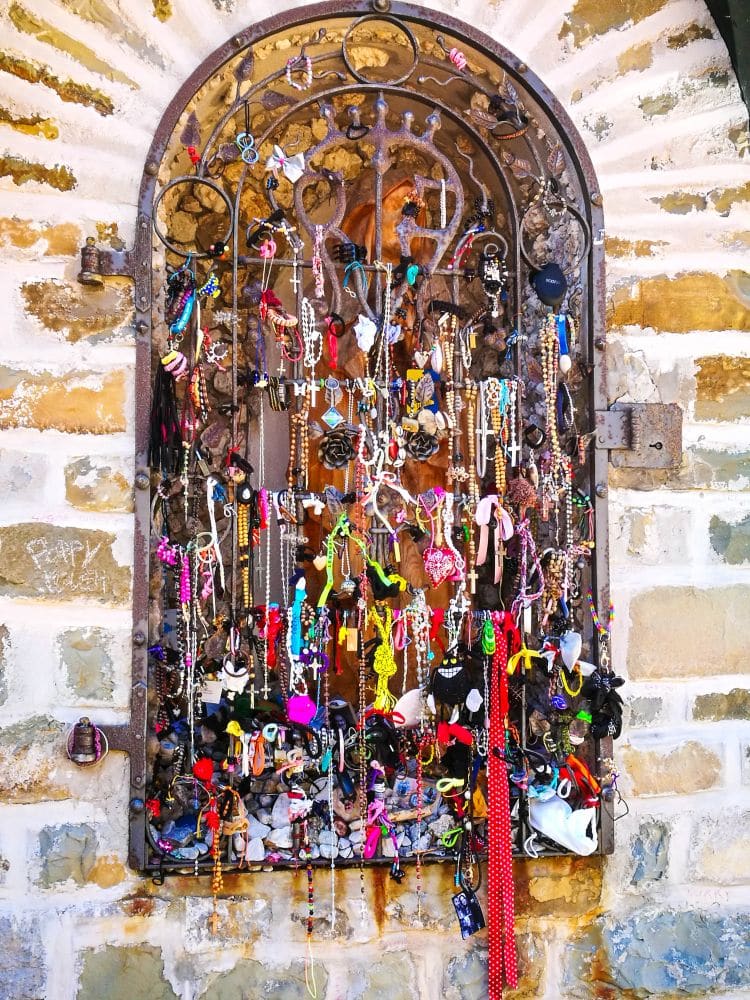 Ein dekoratives eisernes Fenstergitter an einer Steinmauer am Pragser Wildsee, bedeckt mit bunten Bändern, Armbändern, Perlen und Schmuckstücken, die von Besuchern als Opfergaben oder Wünsche hinterlassen wurden.