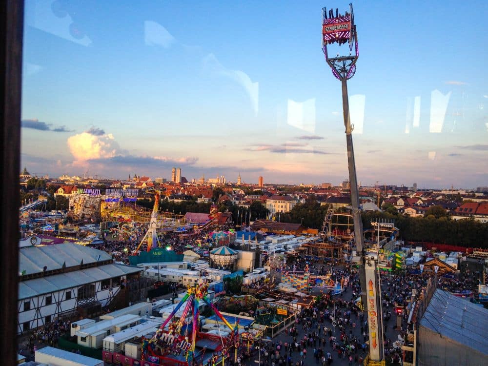 Luftaufnahme eines überfüllten Festplatzes bei Sonnenuntergang während des Oktoberfestes mit bunten Fahrgeschäften, Buden, einer großen Schaukel und der Skyline der Stadt im Hintergrund unter blauem Himmel.