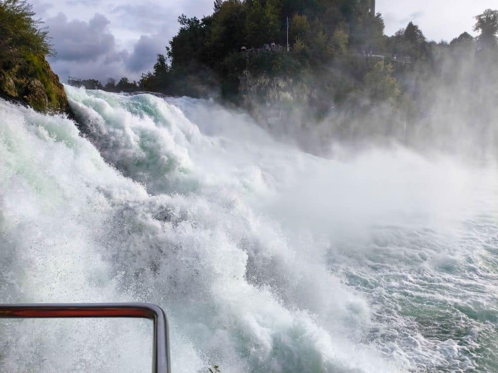 Ein mächtiger Wasserfall stürzt über Felsen und erzeugt Nebel und weißes Wasser, mit üppig grünen Bäumen am Flussufer unter einem bewölkten Himmel. Links unten ist ein rotes Metallgeländer zu sehen - das ist der berühmte Rheinfall von Schaffhausen, Europas größter Wasserfall.