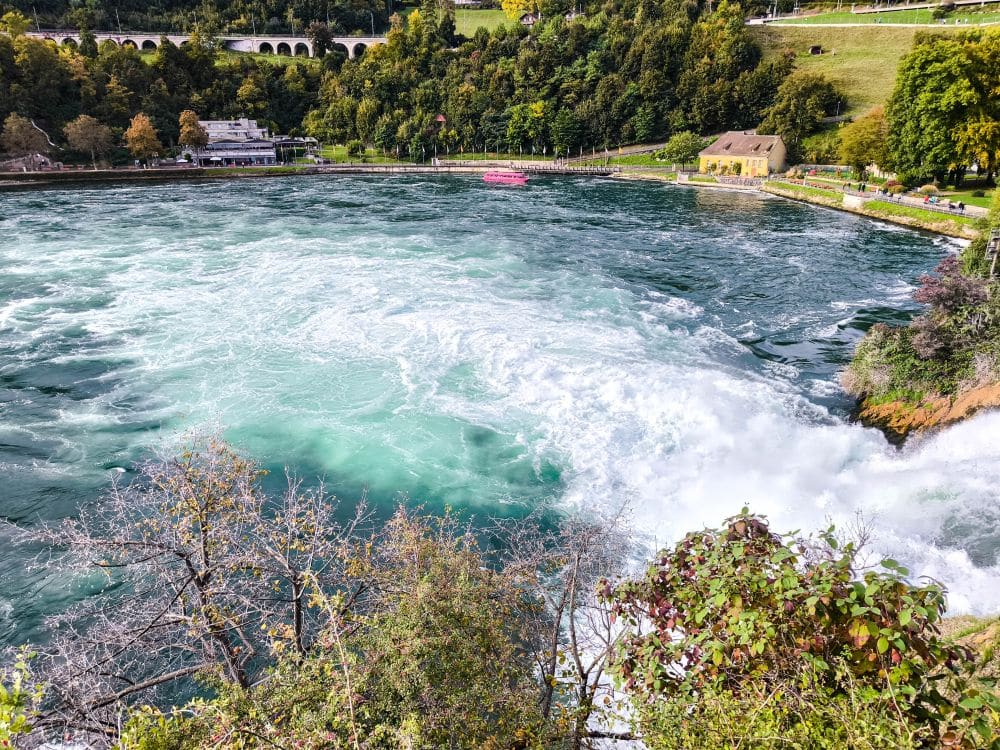 Ein großer, türkisfarbener Wasserstrudel am Fuße des Rheinfalls, Europas größtem Wasserfall, umgeben von grünen Bäumen und Gebäuden, mit einer Eisenbahnbrücke im Hintergrund bei Schaffhausen.