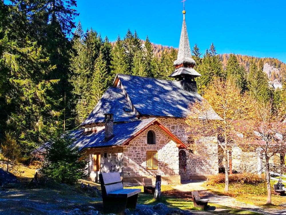 Eine steinerne Kirche mit steilem Schieferdach und spitzem Turm steht unter hohen, immergrünen Bäumen bei strahlend blauem Himmel in der Nähe des Pragser Wildsees, mit einer Bank und Herbstlaub im Vordergrund.