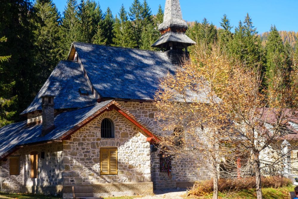 In der Nähe des Pragser Wildsees steht zwischen herbstlichen Bäumen und immergrünen Bäumen eine kleine Steinkirche mit Holzdach und Kirchturm, auf deren Mauern das Sonnenlicht Schatten wirft.