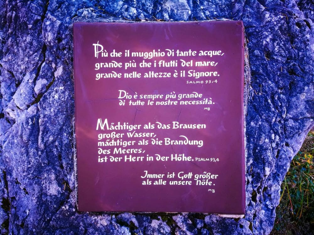 Eine violette Tafel mit religiösem Text in italienischer und deutscher Sprache, einschließlich Bibelversen, ist auf einer felsigen Oberfläche in der Nähe des Pragser Wildsees angebracht. Der Hintergrund besteht aus strukturiertem Stein und etwas Grün an den Rändern.