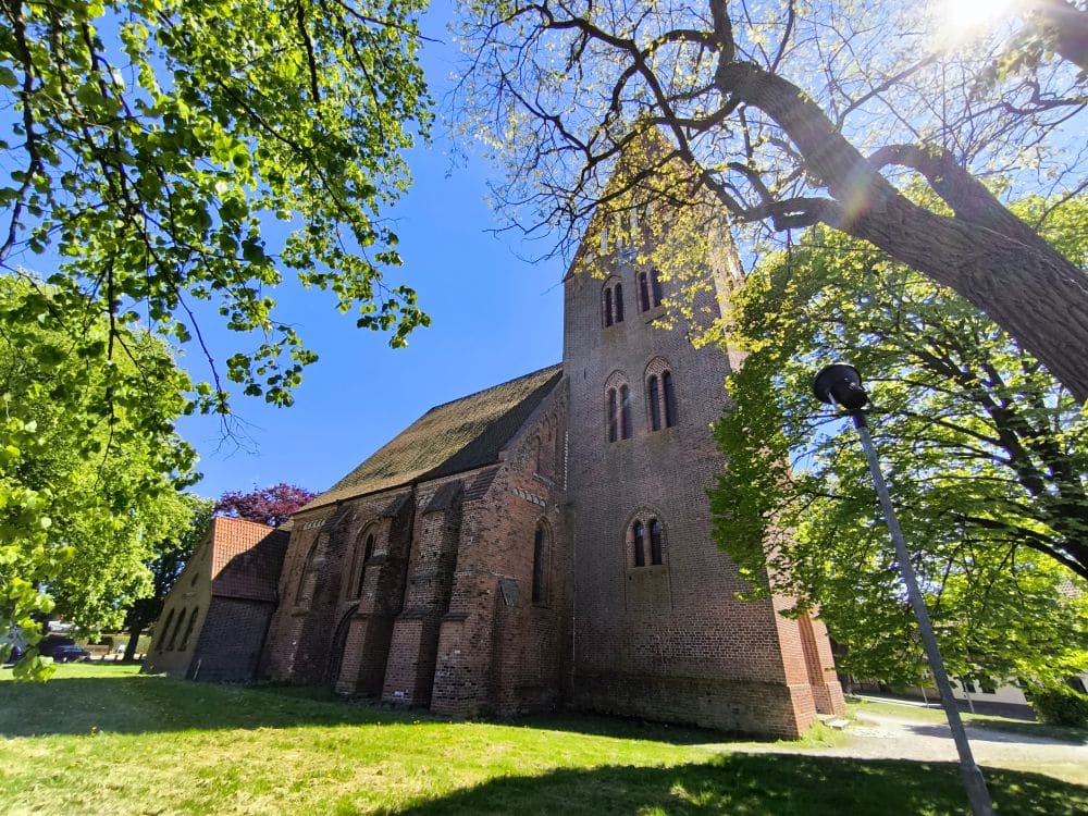 In Klütz, einem versteckten Juwel an der Ostseeküste, steht unter strahlend blauem Himmel eine Backsteinkirche mit Bogenfenstern und einem hohen Turm, umgeben von grünen Bäumen, durch deren Äste das Sonnenlicht fällt.