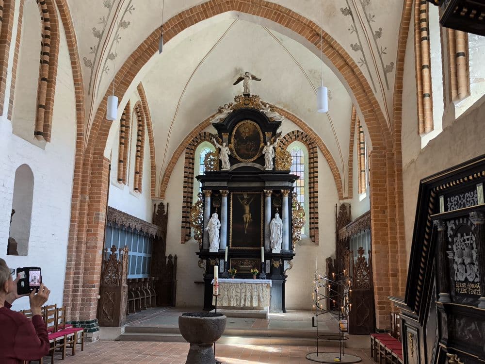 Eine Person macht ein Foto in einer Kirche in Klütz an der Ostseeküste mit gewölbten Decken, weißen Backsteinwänden, hölzernen Kirchenbänken und einem reich verzierten Altar mit Statuen und goldenen Akzenten, der durch das natürliche Licht aus hohen Fenstern beleuchtet wird.