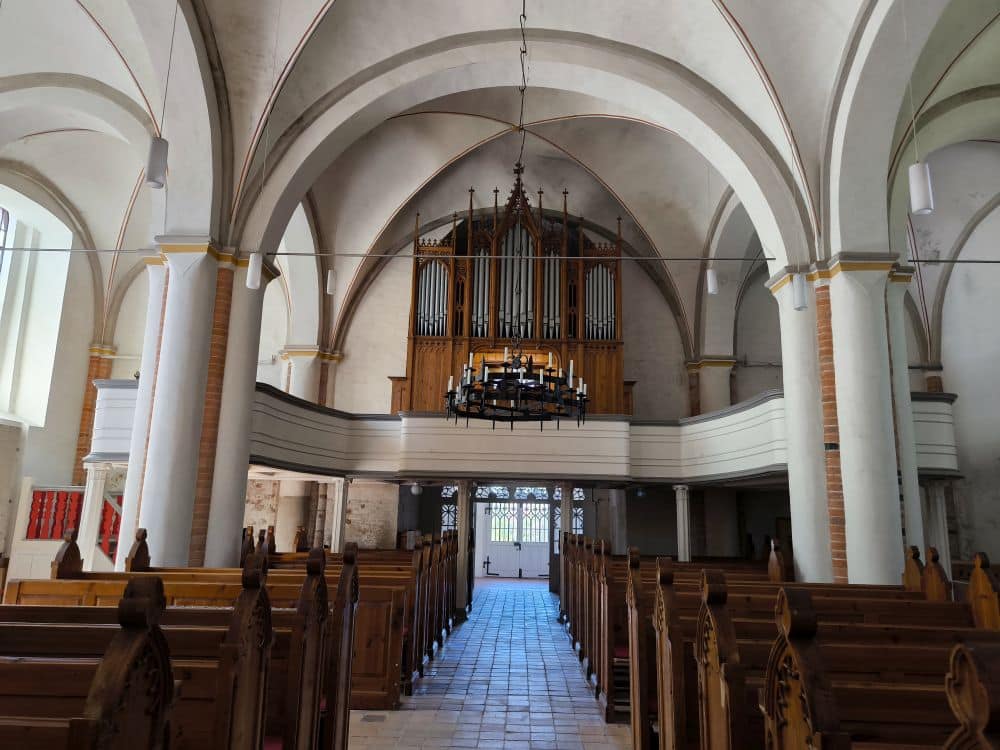 Innenraum einer Kirche in Klütz an der Ostseeküste mit Holzbänken, die einen Mittelgang flankieren, der zu einer großen Pfeifenorgel führt. Gewölbte Decken, stattliche Säulen und ein kreisförmiger Kronleuchter tragen zu der heiteren Atmosphäre bei.
