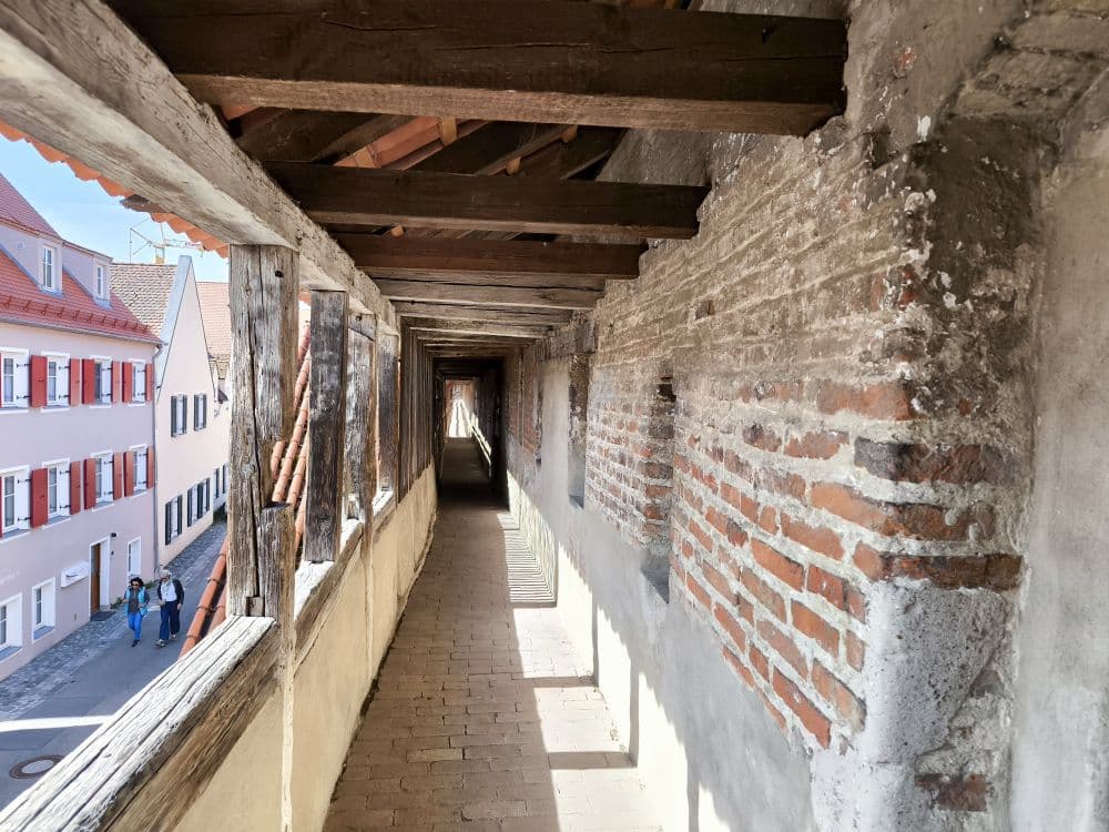 Blick aus dem Inneren eines überdachten Gehwegs aus Ziegeln und Holz auf der Nördlinger Stadtmauer auf den Korridor mit einfallendem Sonnenlicht. Unten gehen zwei Personen neben bunten Gebäuden entlang - perfekt für eine Entdeckungstour in der Kraterstadt.