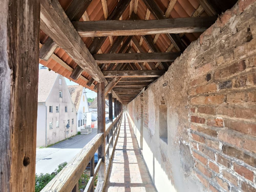 Überdachter Holzsteg mit Ziegeldach, der entlang einer alten Backsteinmauer verläuft, ideal für eine Entdeckungstour durch die historische Kraterstadt Nördlingen, mit Blick auf eine ruhige Straße und charmante Häuser im Tageslicht.