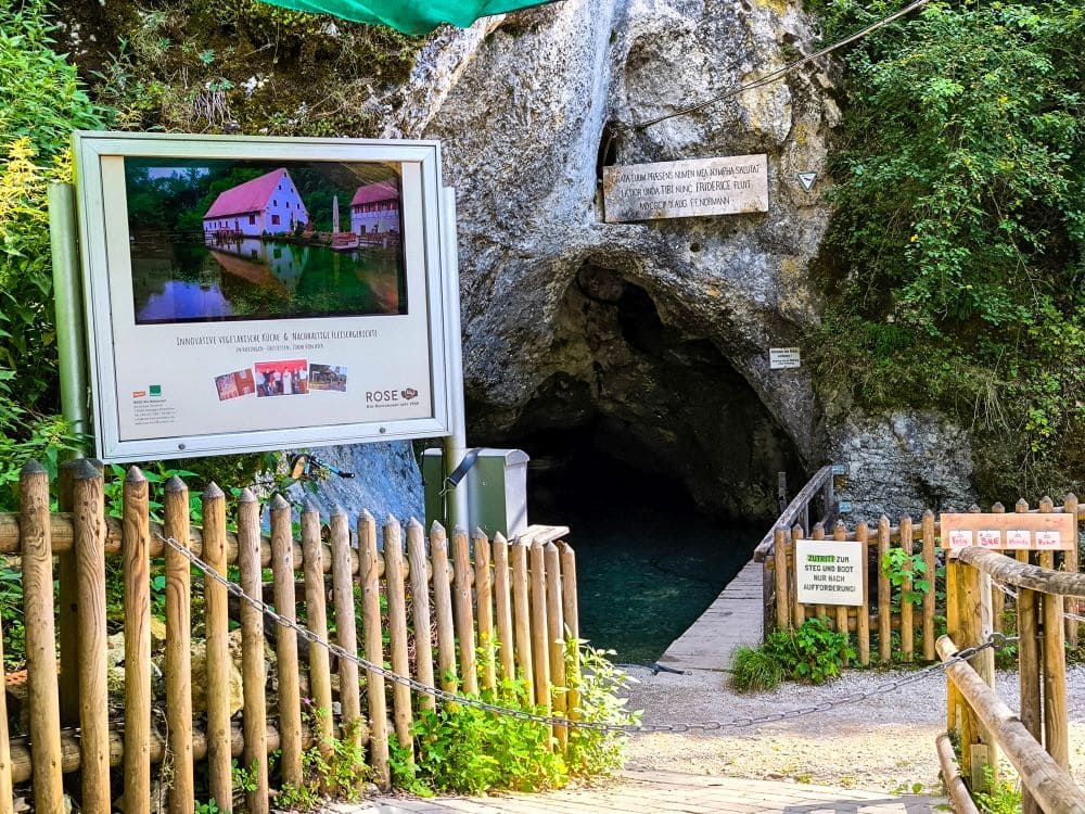 Ein Höhleneingang bei Hayingen, umgeben von Grün und einem Holzzaun, empfängt den Wanderer. Auf der linken Seite befindet sich eine große Informationstafel mit einem Hausfoto, und eine kleine Holzbrücke führt zur Höhlenöffnung der Wanderung Glastal.