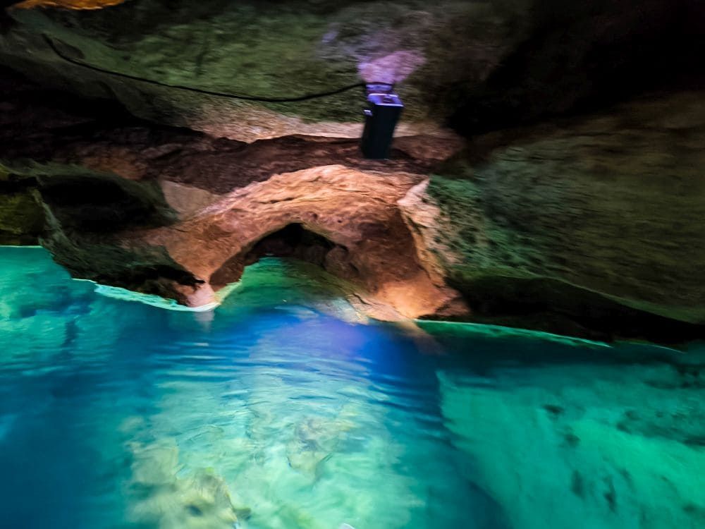 Eine Felsenhöhle in der Nähe von Hayingen mit türkisblauem Wasser, das von natürlichem und künstlichem Licht beleuchtet wird, offenbart ein ruhiges unterirdisches Becken und eine strukturierte Steindecke - perfekt, um sie während Ihrer Wanderung Glastal oder Hochgehschätzt zu erkunden.