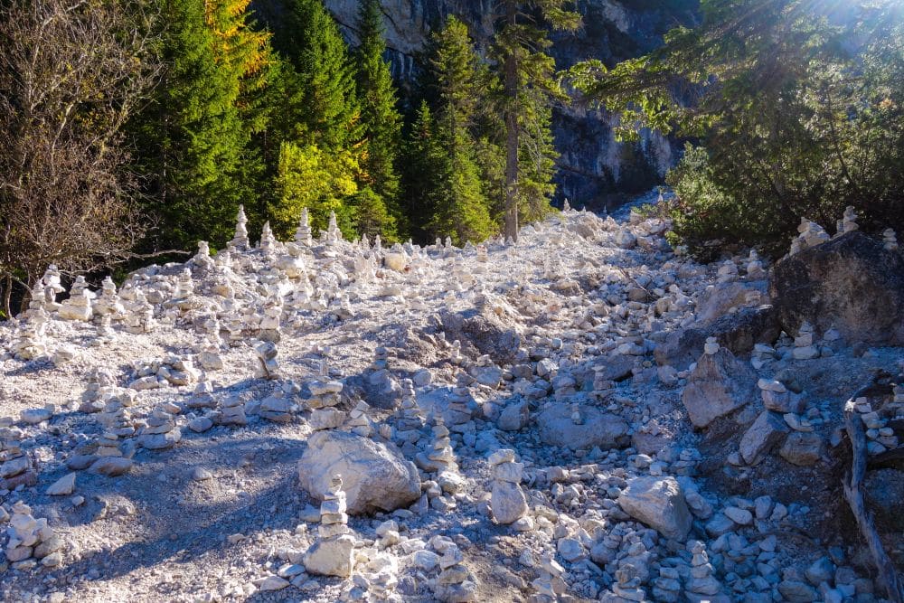 Eine felsige Waldlichtung in der Nähe des Pragser Wildsees, gefüllt mit kleinen Stapeln weißer Steine im Sonnenlicht, umgeben von grünen Bäumen und vor der Kulisse hoch aufragender Felsen.