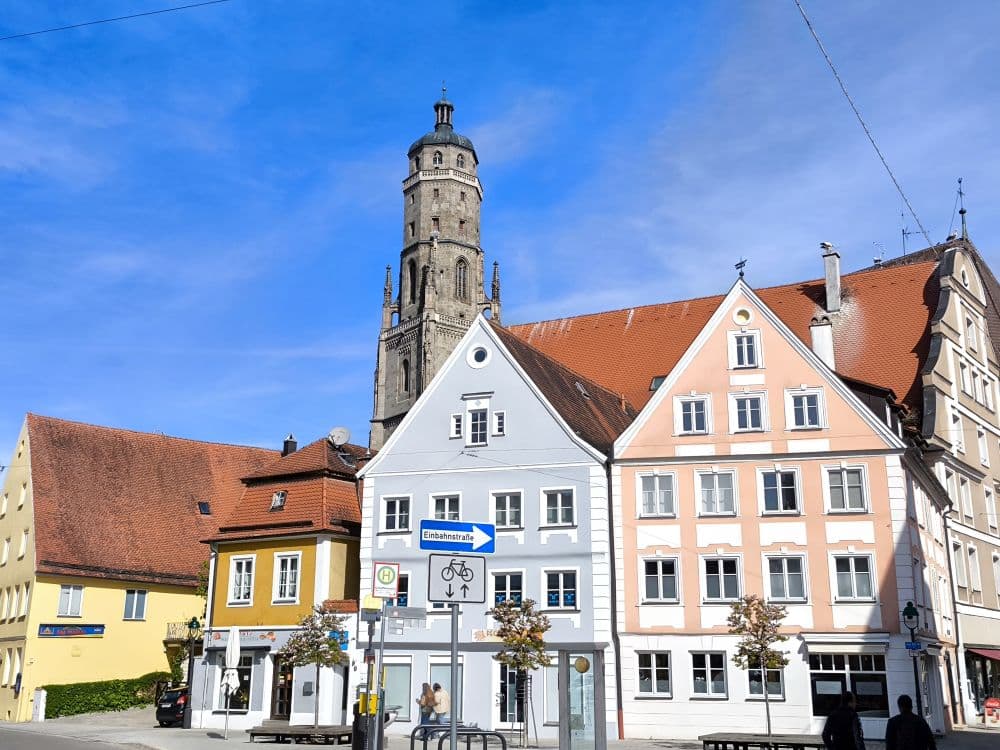 Eine Straßenszene in Nördlingen zeigt bunte Gebäude mit steilen Dächern und weiß gerahmten Fenstern, die von einem hohen steinernen Kirchturm im Hintergrund unter einem strahlend blauen Himmel dominiert werden.