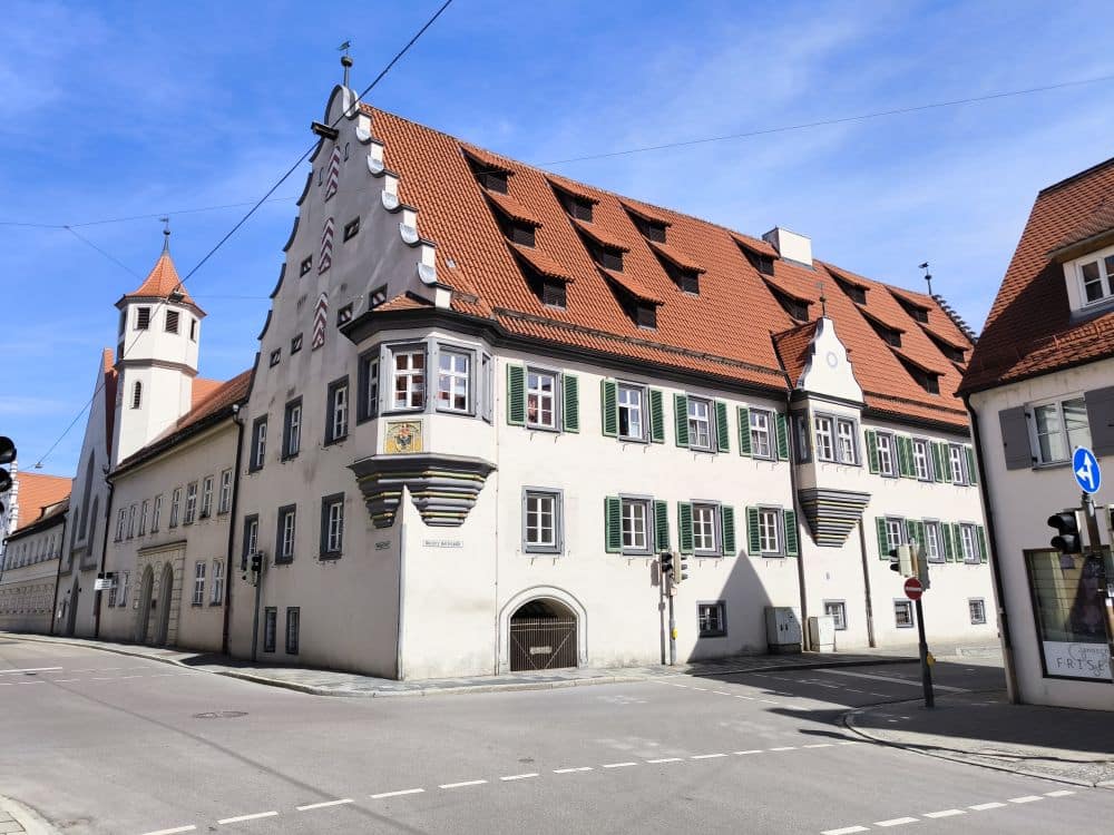 Ein historisches weißes Gebäude mit einem steilen roten Ziegeldach, Dachgauben, grünen Fensterläden und dekorativen Verzierungen steht an einer sonnigen Straßenecke in Nördlingen, einer charmanten europäischen Stadt. Ein blauer Himmel ist über dem Gebäude zu sehen.