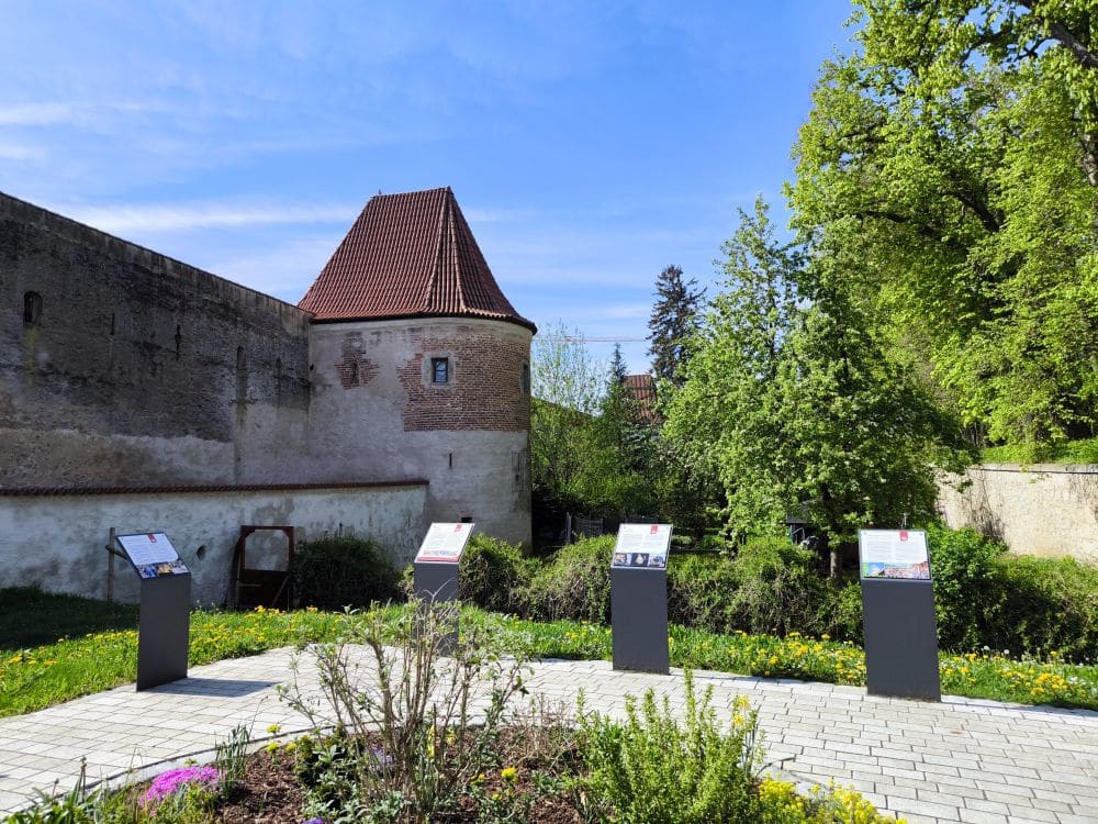Eine historische Steinmauer mit einem runden, rot gedeckten Turm in Nördlingen steht neben vier Informationstafeln an einem gepflasterten Weg, umgeben von grünen Bäumen, Gras und einem kleinen Blumenbeet unter blauem Himmel.