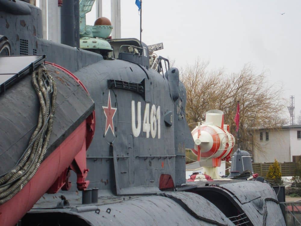 Nahaufnahme eines grauen U-Boots mit der Kennzeichnung U461 und einem roten Stern, das im Freien mit Seilen auf dem Deck angedockt ist. Im Hintergrund ist zwischen Bäumen und Gebäuden ein weiß-orangefarbenes Peenemünde-Torpedo oder -Tauchboot zu sehen.