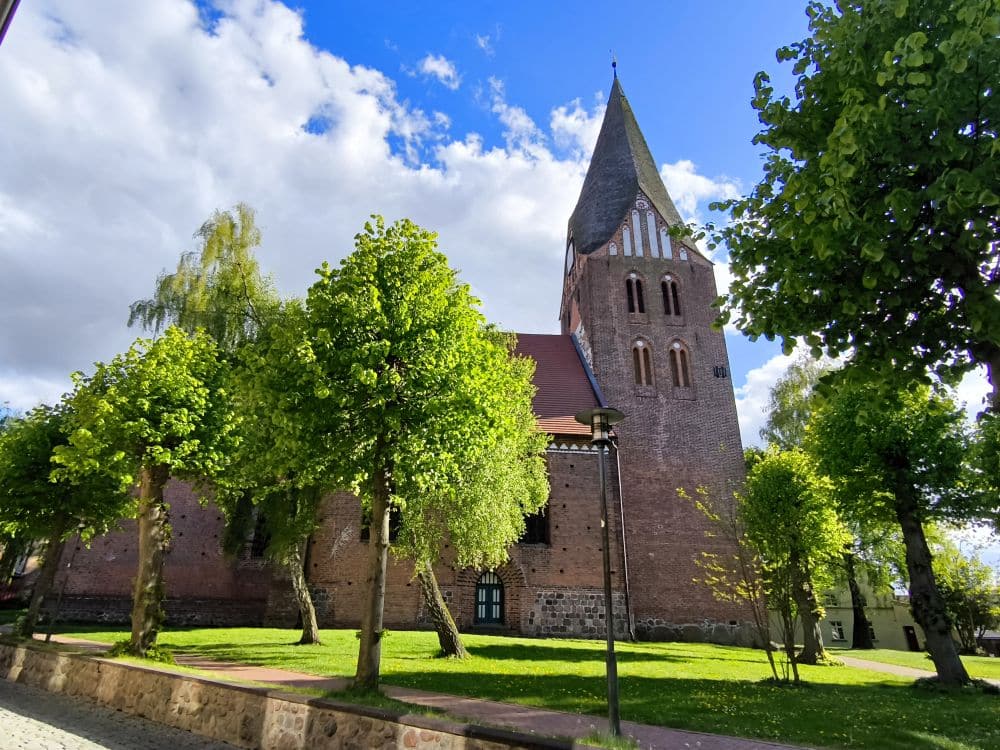 Eine historische Backsteinkirche in Neubukow mit einem hohen, spitzen Kirchturm steht inmitten grüner Bäume unter einem teilweise bewölkten blauen Himmel. Das Sonnenlicht wirft Schatten auf das Gras und das Gebäude und schafft eine friedliche Atmosphäre.