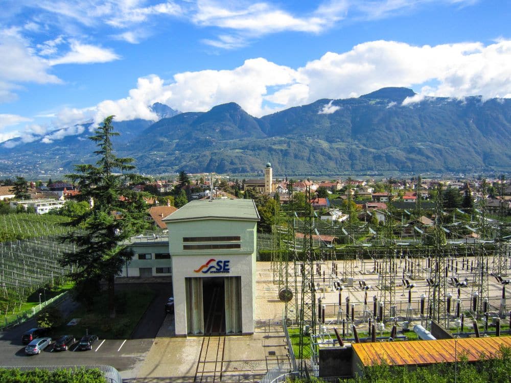 Ein kleines Kraftwerk mit dem SE-Logo befindet sich am Brandiswaalweg vor einem eingezäunten Umspannwerk, umgeben von grünen Feldern und Bäumen, mit einer Stadt und Bergen unter einem teilweise bewölkten Himmel im Hintergrund.